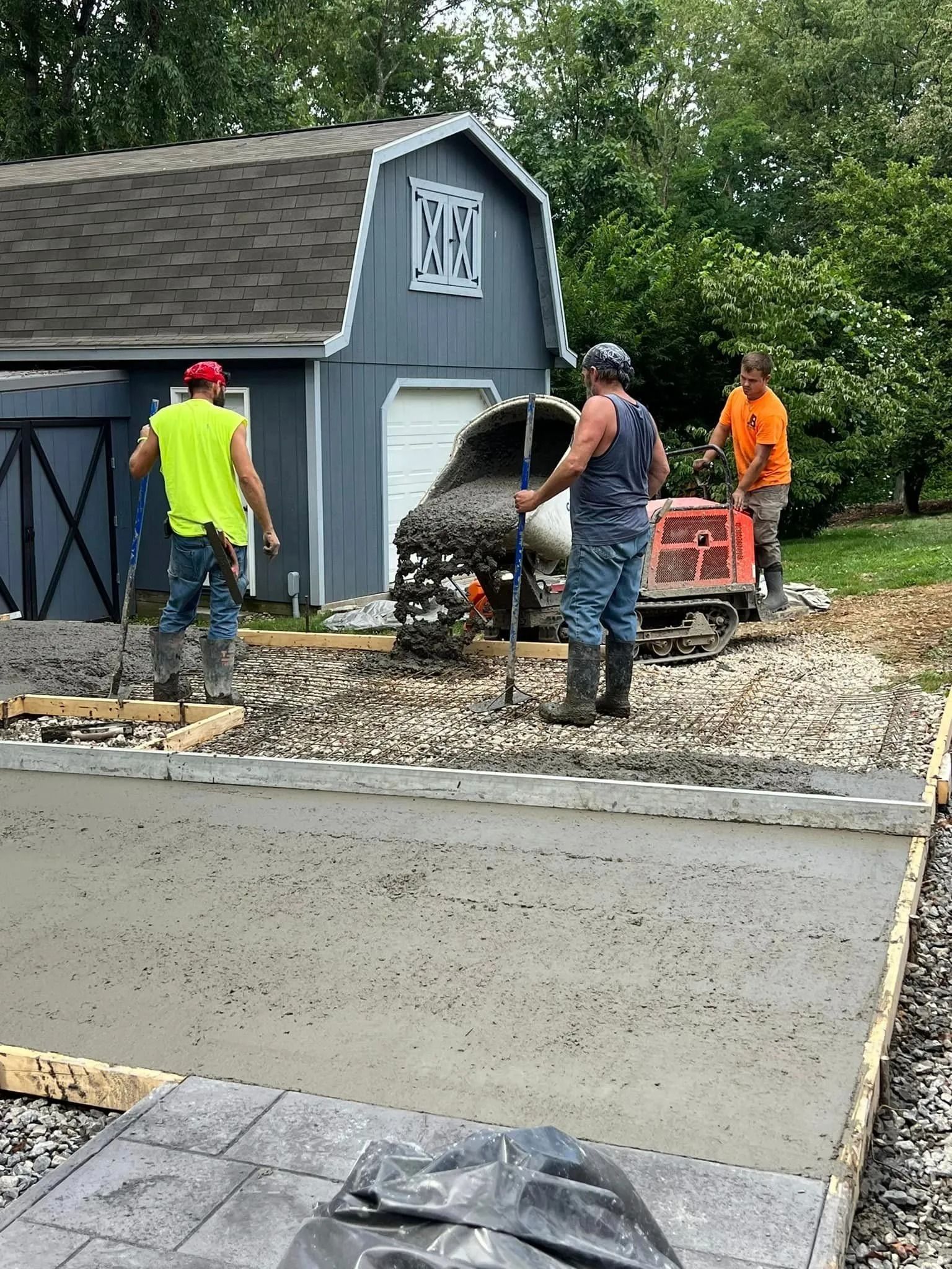 Construction workers pouring concrete for a patio near a blue shed, using a concrete mixer.