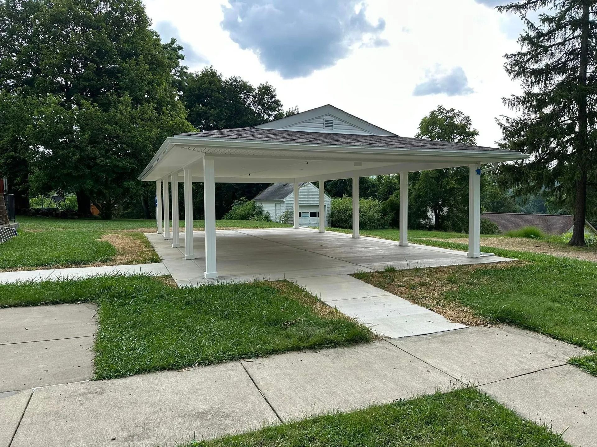 White carport with concrete pillars, on a patch of grass. A sidewalk in foreground.