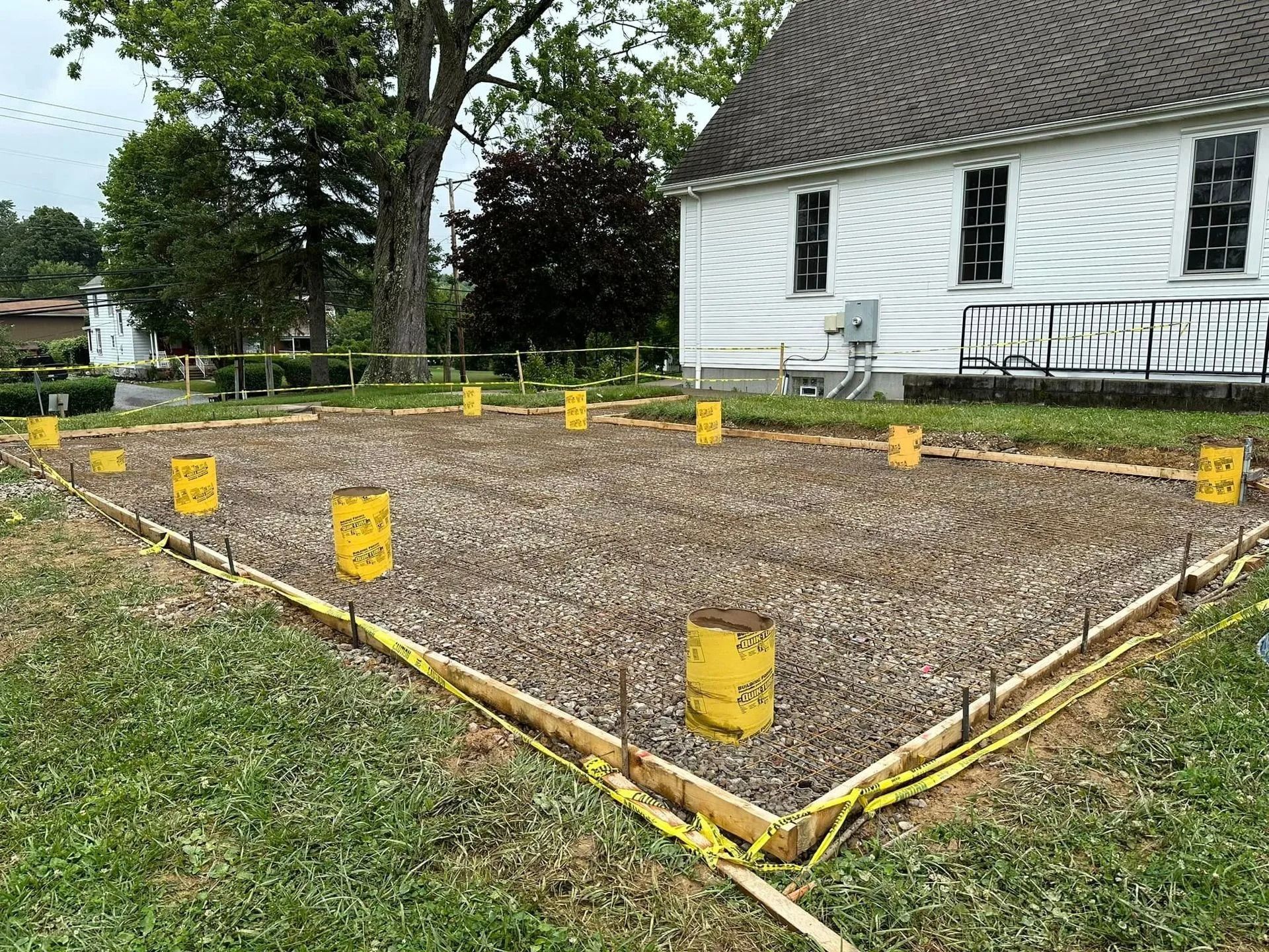 Construction site with wooden frame and gravel base, yellow markers, and a white building in background.