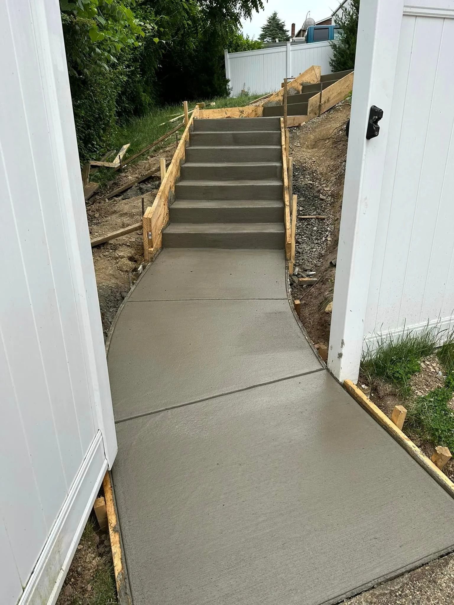 Concrete walkway leading to concrete steps up a small hill, flanked by wood and a white fence.