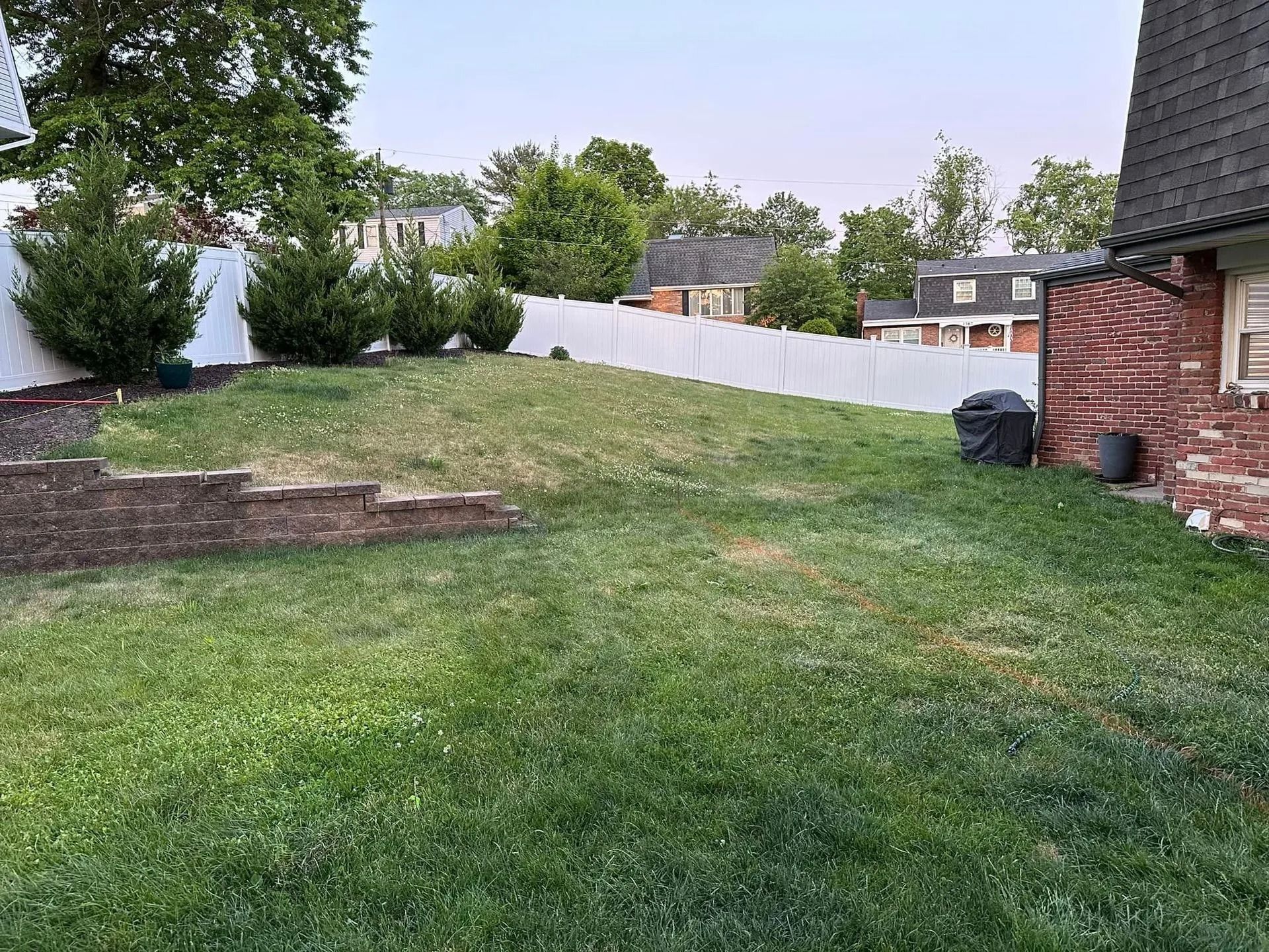 Grassy backyard with a small hill, brick wall, white fence, and part of a house.