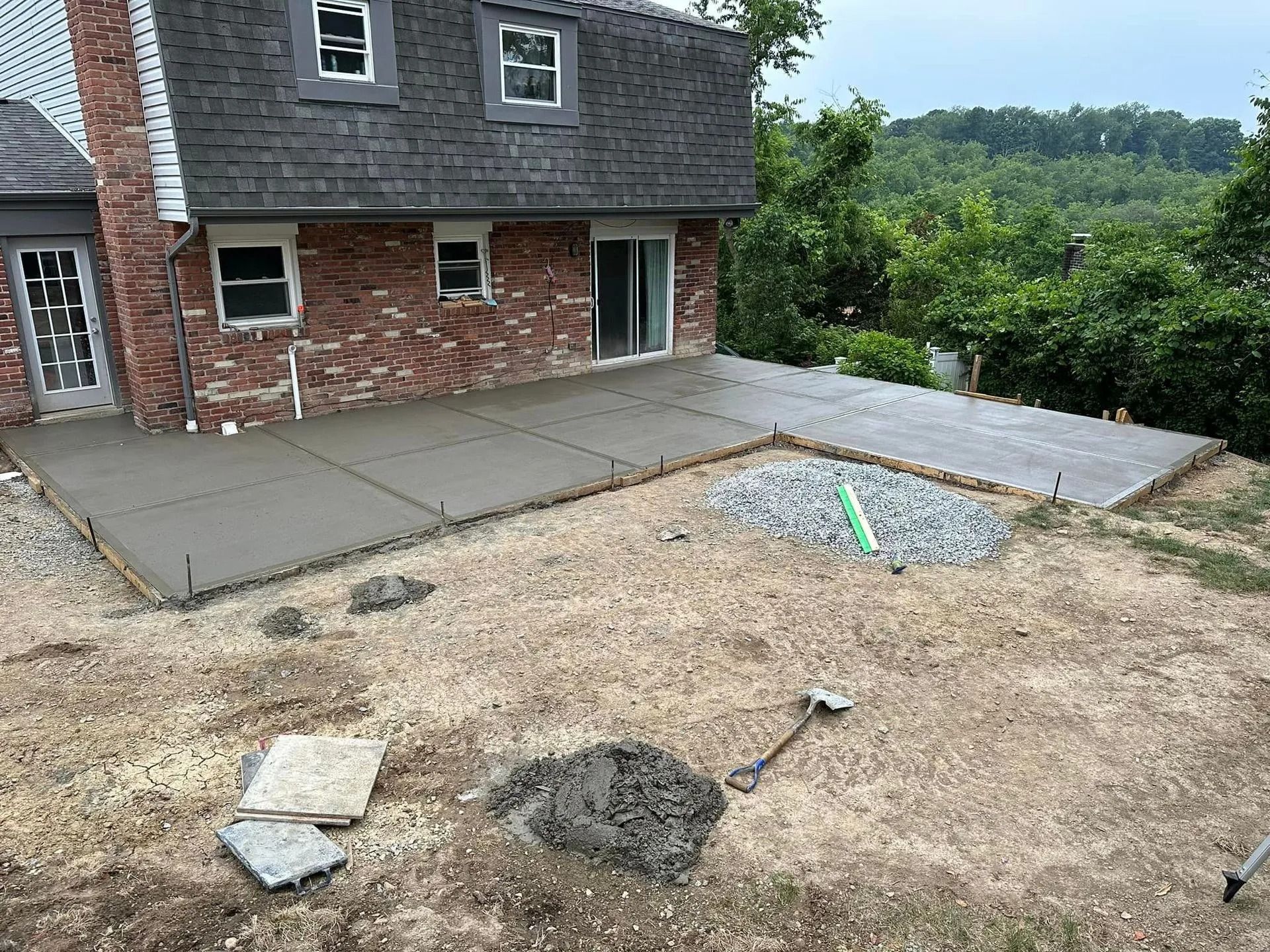 Concrete patio being built next to a brick house with a hillside in the background.