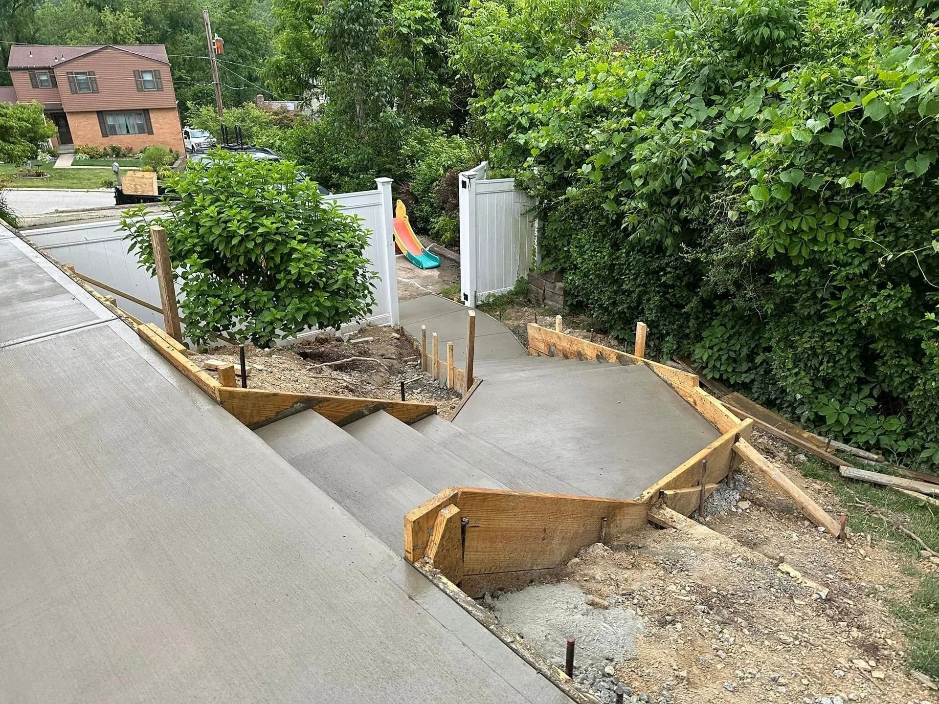 Newly poured concrete steps leading down a slope, framed by wooden forms, beside a house and yard.
