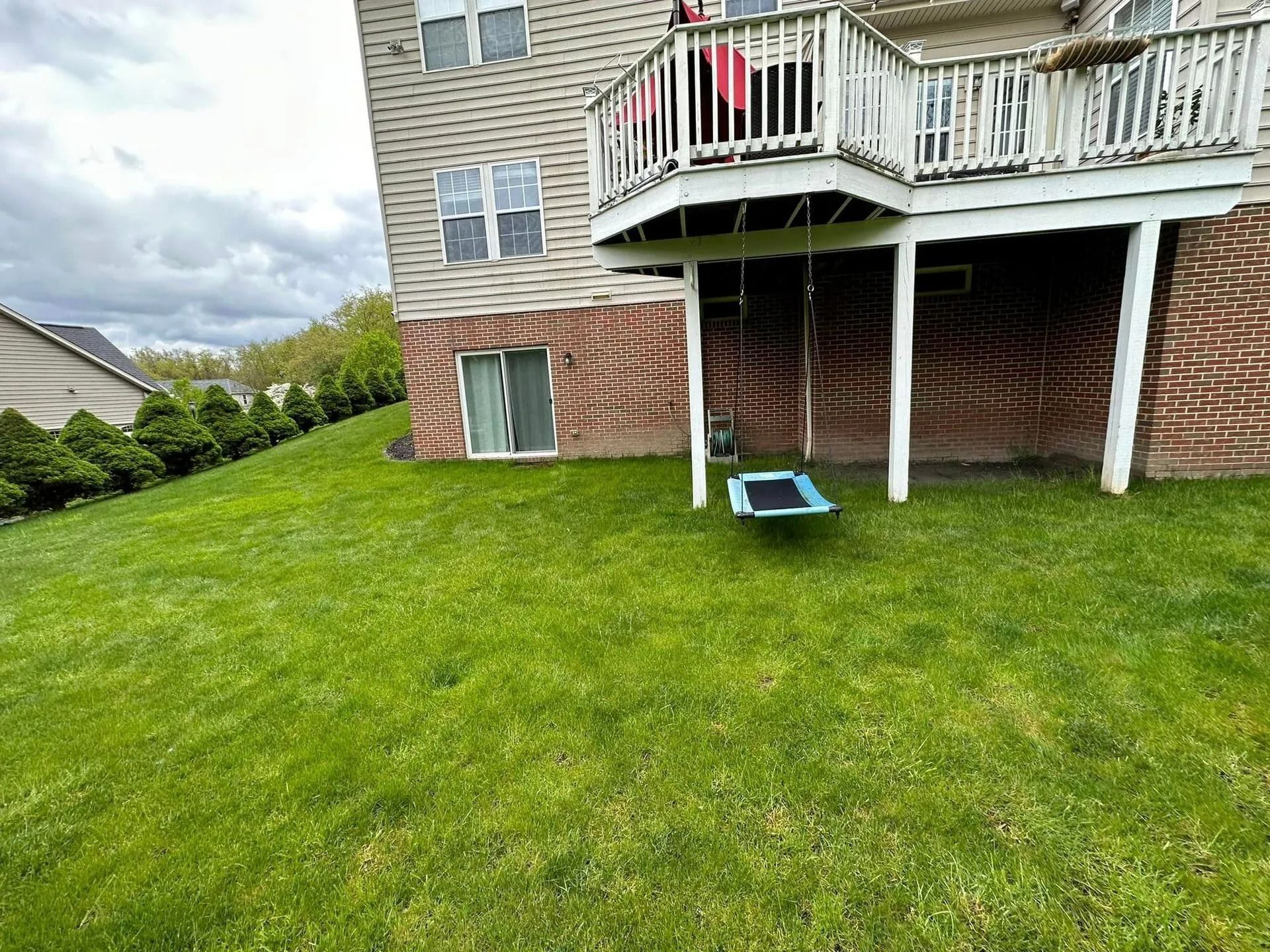 Green lawn slopes up to a two-story house with a deck. A swing hangs below the deck. Overcast sky.