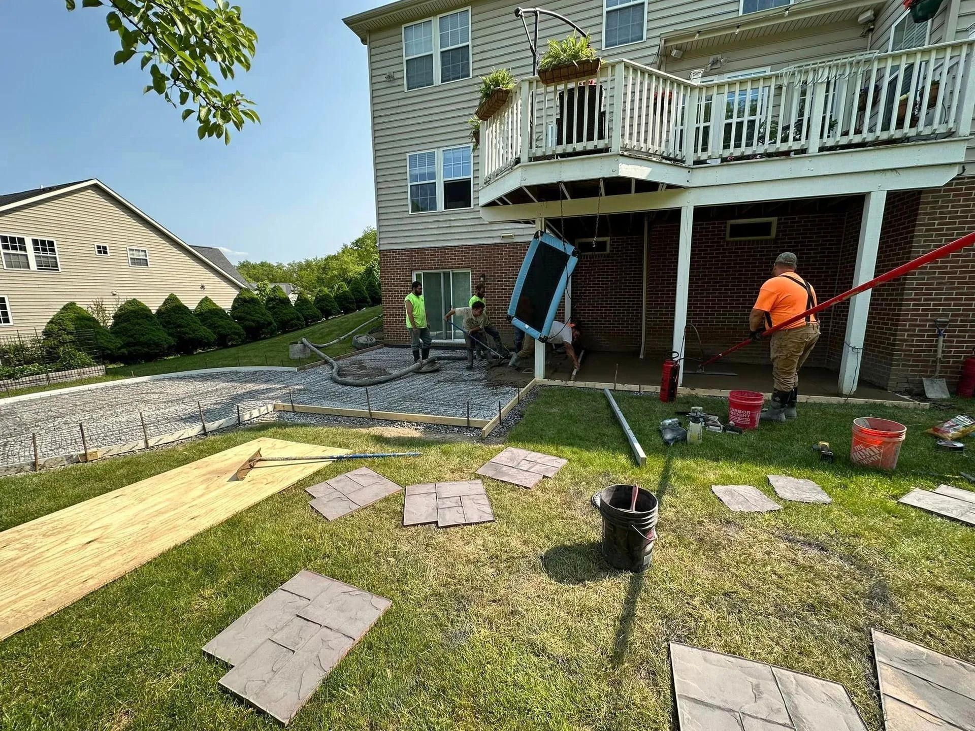 Construction workers installing patio stones and working under a deck, near a two-story house.