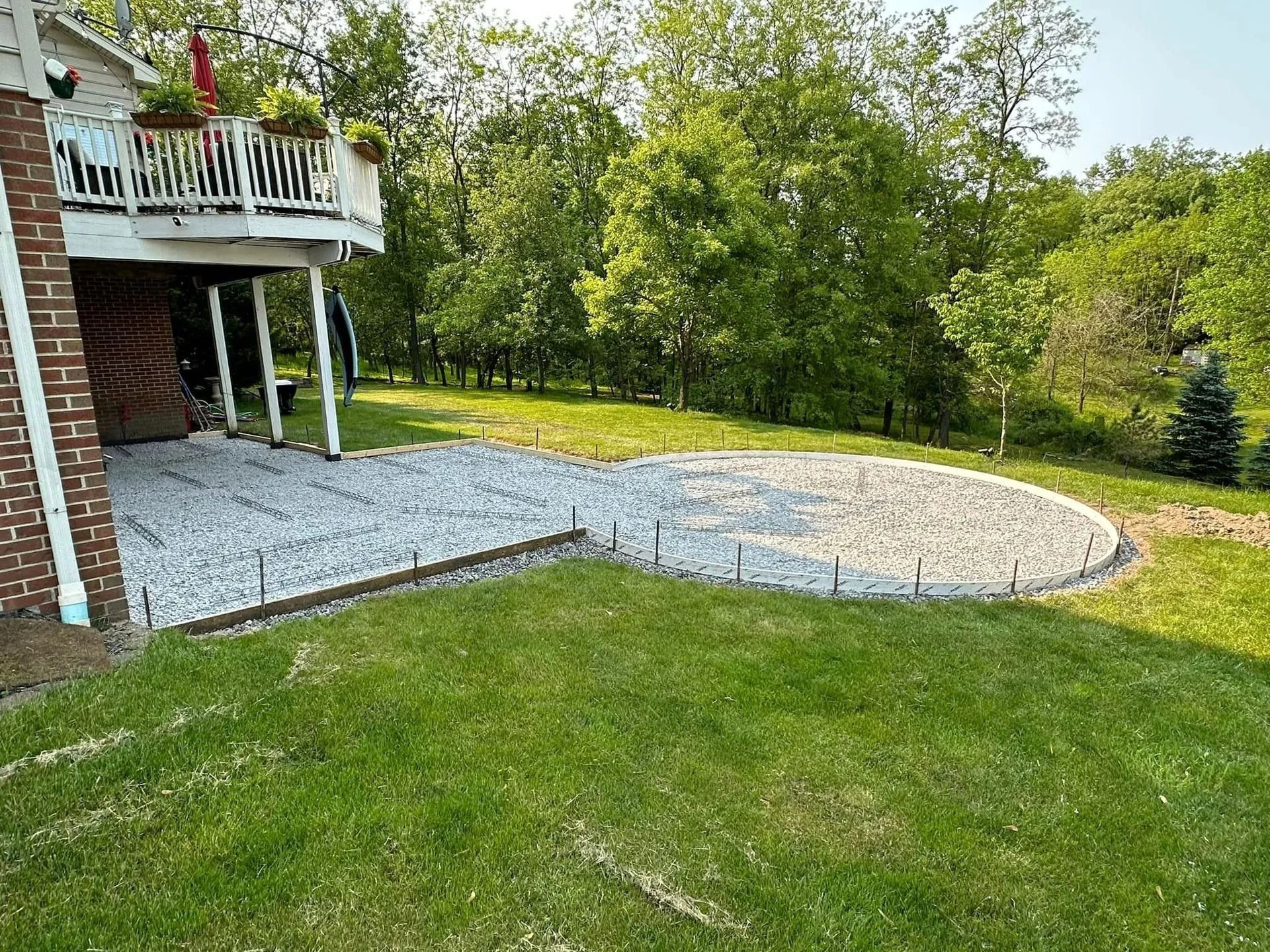 A backyard with a gravel patio under a deck, surrounded by grass and trees.