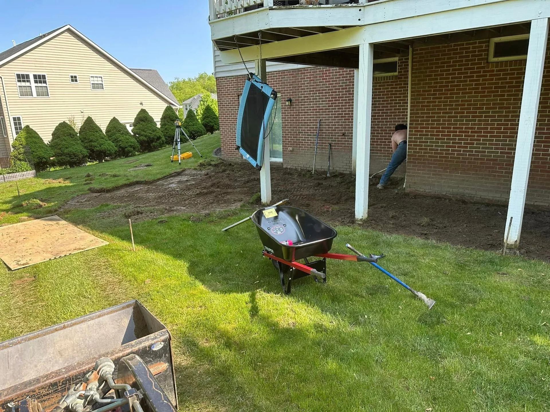 Backyard landscaping project in progress: dirt, tools, wheelbarrow, and a person working under a deck.