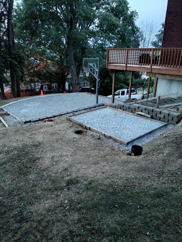 Gravel basketball court under construction in a yard, bordered by wooden beams. A deck overlooks the area.