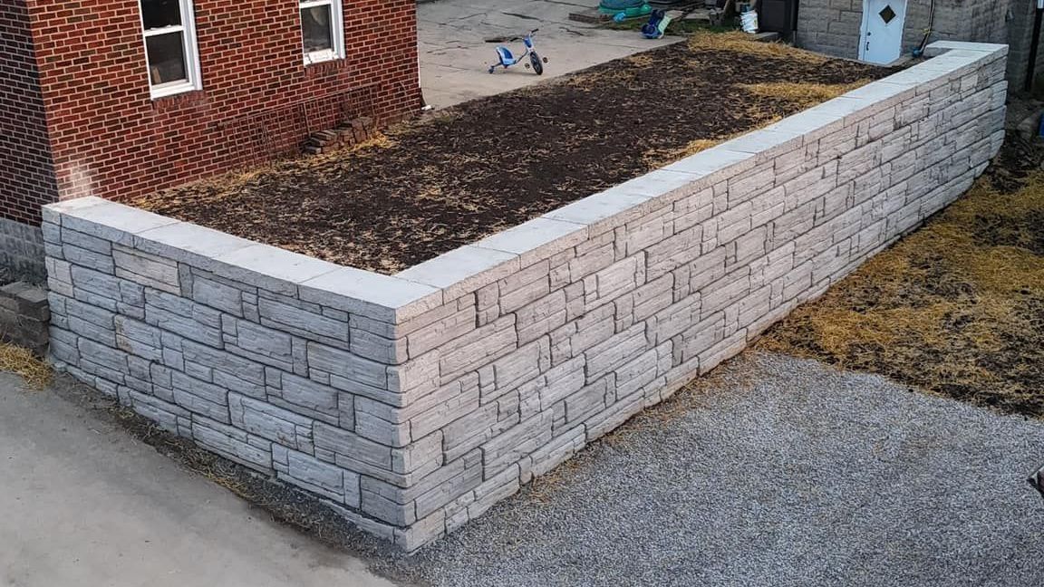 Retaining wall made of gray concrete blocks beside a brick building and a gravel driveway.