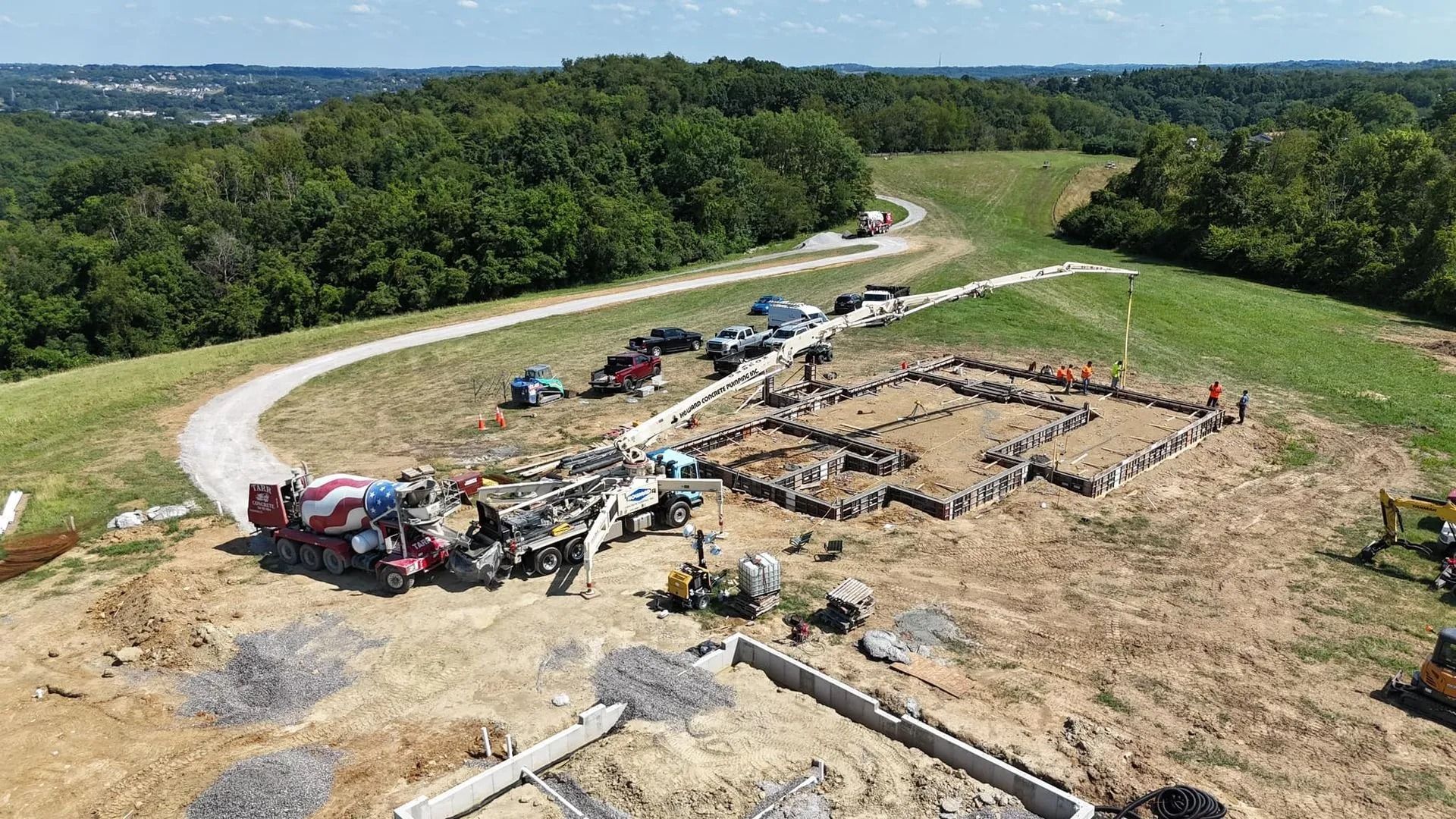 Construction site with cement truck pouring concrete into a foundation. Workers are present; the landscape is green and hilly.