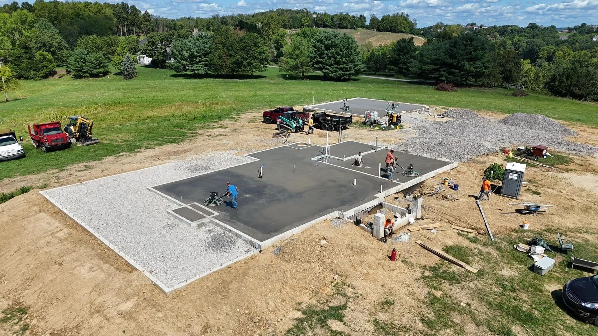 Construction site with concrete foundation and workers on a grassy hill; trucks and equipment present.