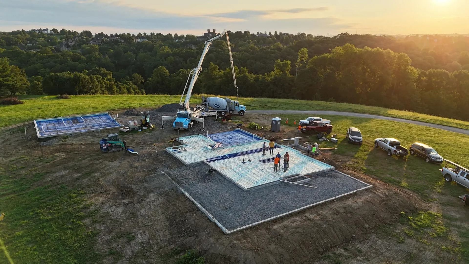 Construction site with concrete being poured for a foundation. Blue truck with a boom, workers, and a grassy hilltop setting.