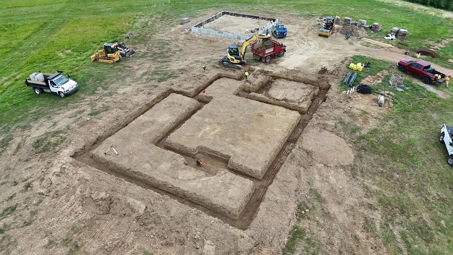 Construction site: excavated foundation with machinery and trucks on a grassy field.