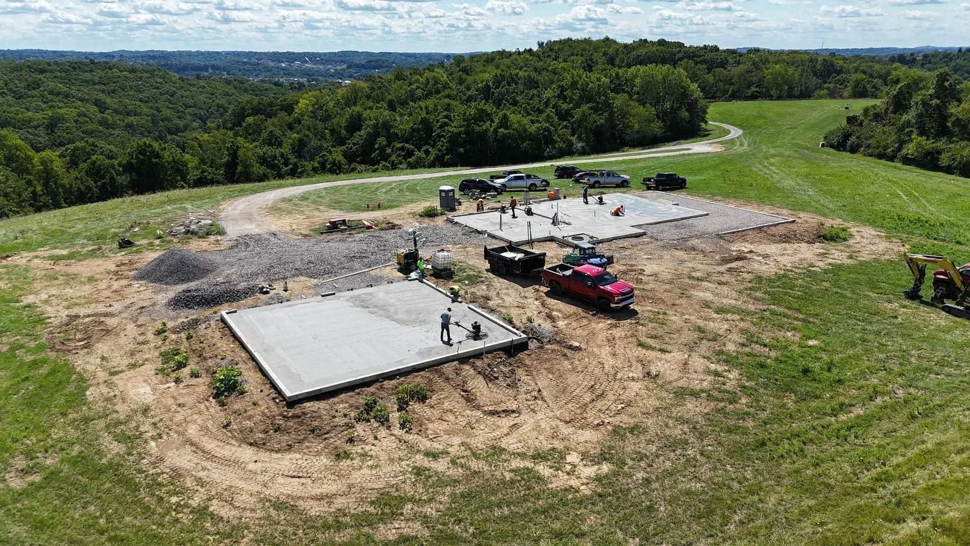 Construction site: Concrete foundations on a grassy hill, surrounded by trees and a red truck.
