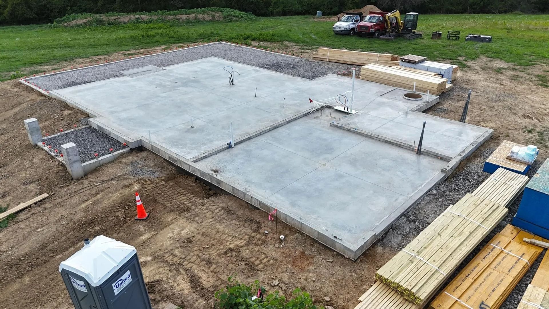 Aerial view of a concrete foundation for a building, with exposed footers and plumbing, surrounded by dirt and lumber.