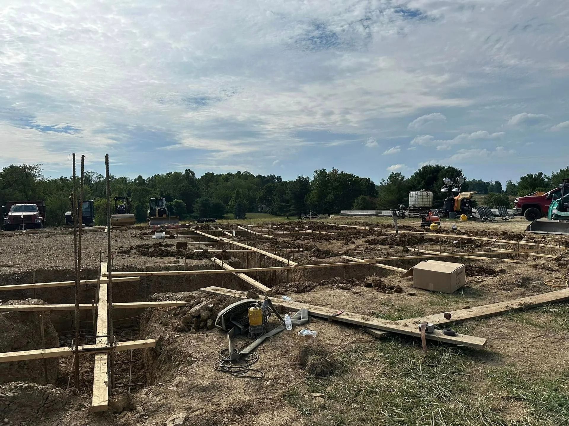 Construction site with wooden forms for foundation; dirt ground; trees and sky in background.