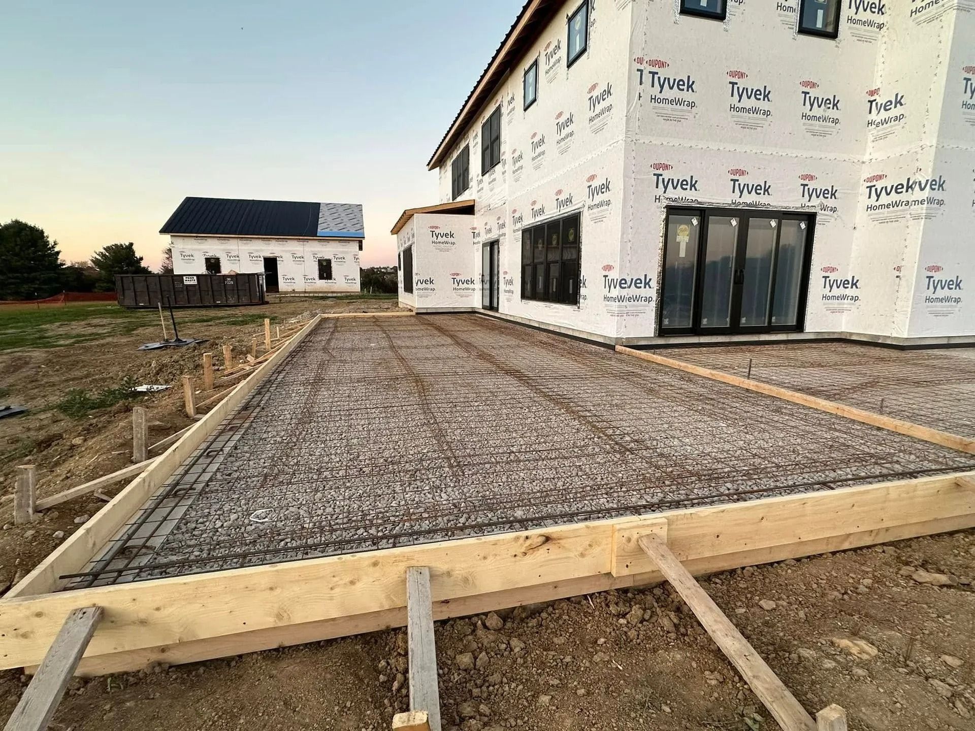 Construction site: Concrete foundation framework with rebar, next to a partially built house wrapped in Tyvek.
