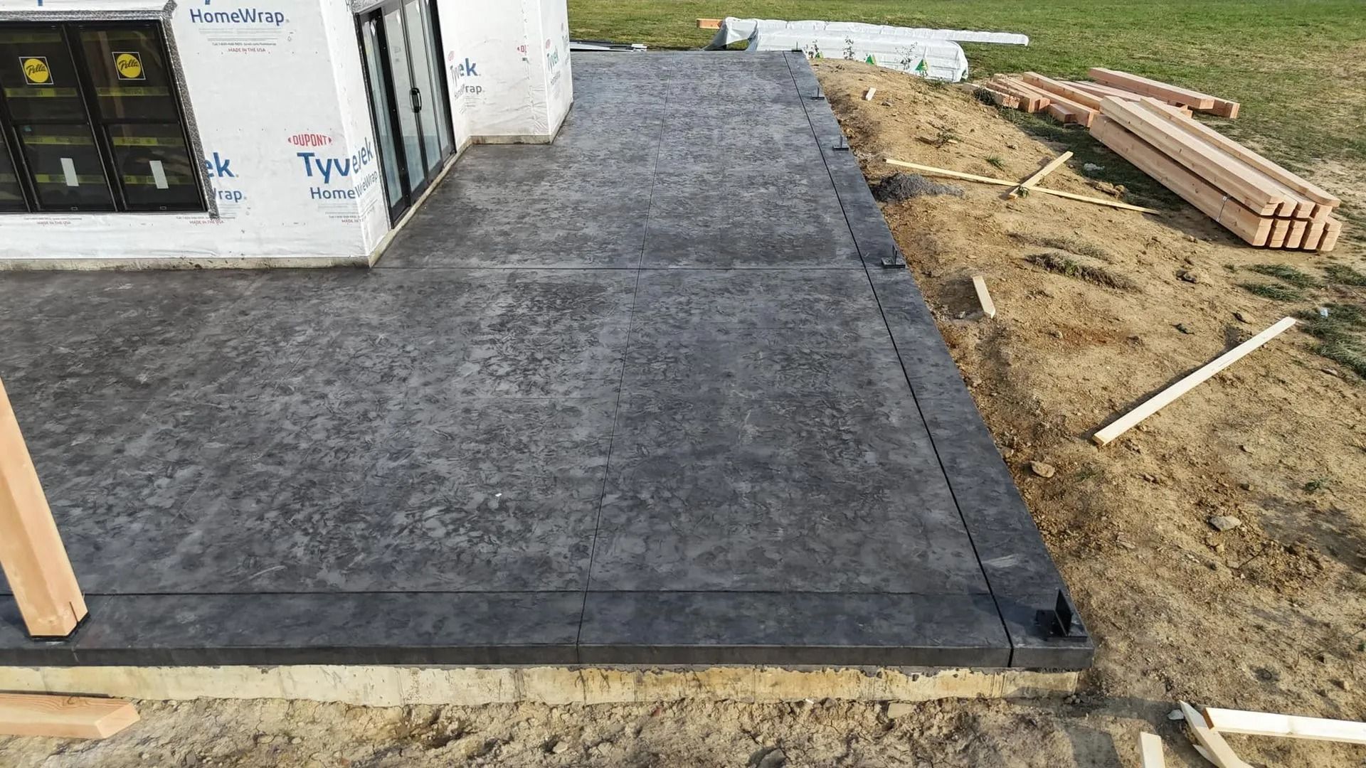 Gray concrete patio beside a building under construction, with wood beams and dirt visible.