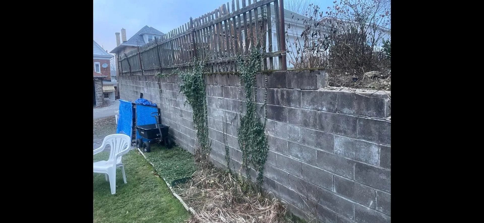 A gray concrete block wall with wooden fence on top and ivy growing on the wall, a lawn and a white chair in the foreground.