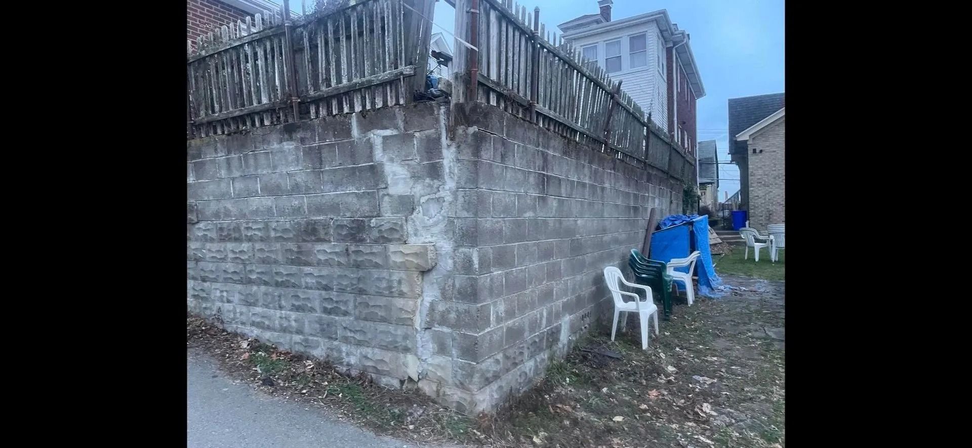 A weathered brick wall with a wooden fence. White chairs and tarp in a backyard.