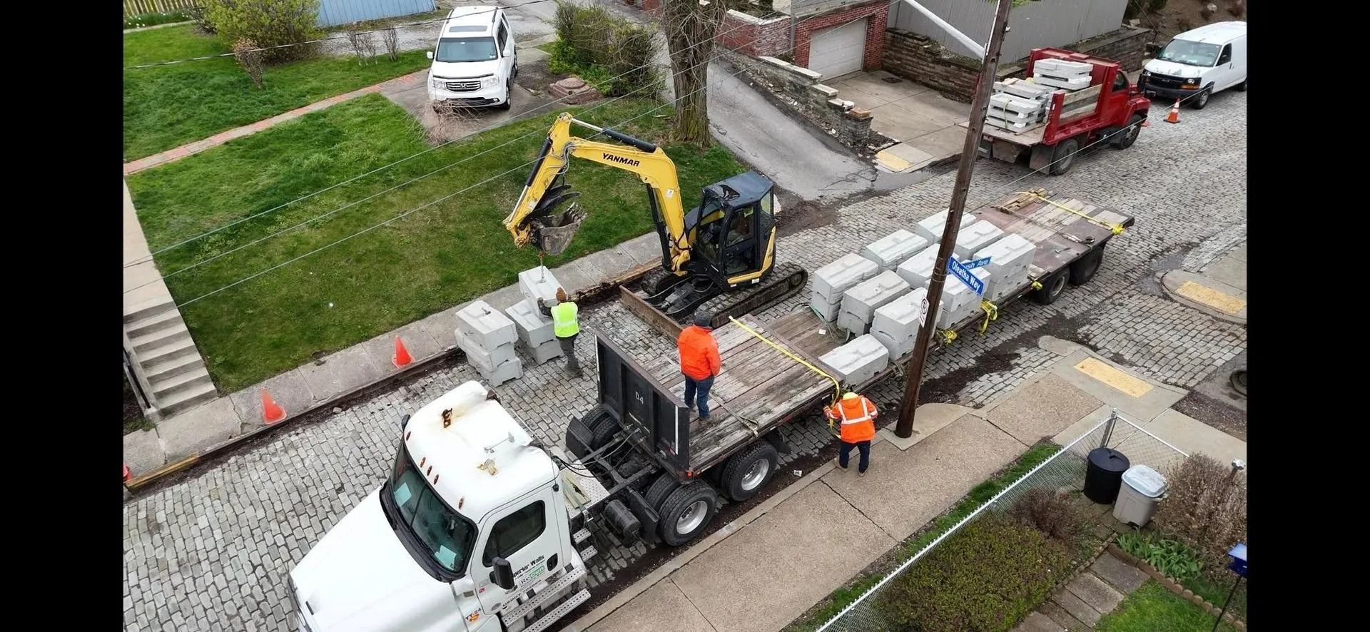 Construction workers unloading concrete blocks with an excavator from a flatbed truck on a cobblestone street.