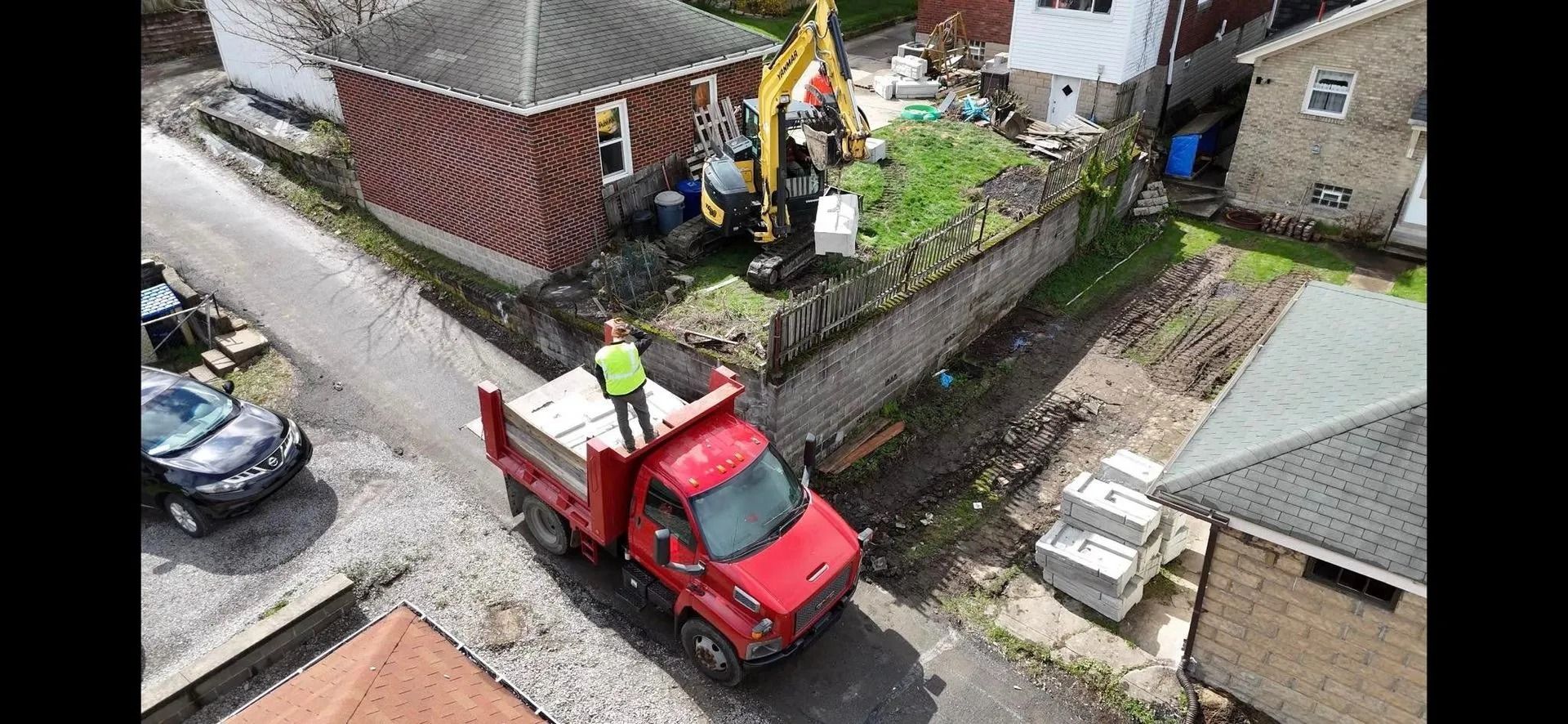 Construction site with a red truck, excavator, and worker standing on the truck bed, next to a brick house.