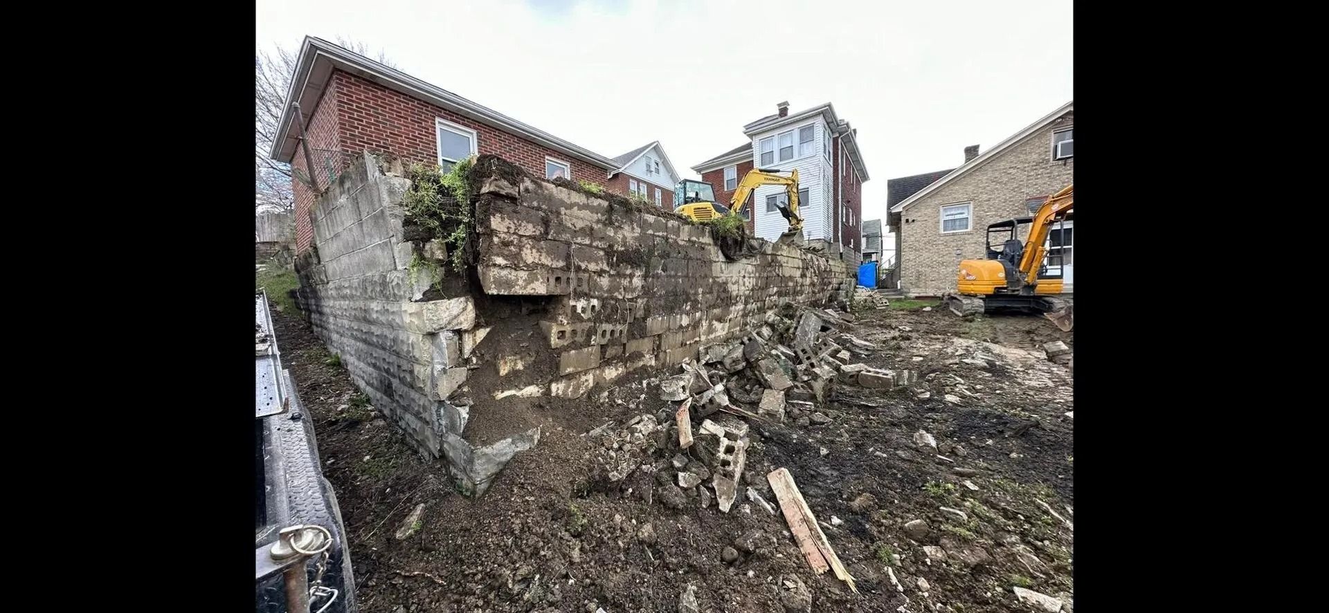 Construction site with excavators demolishing a stone wall. Debris and buildings in the background.