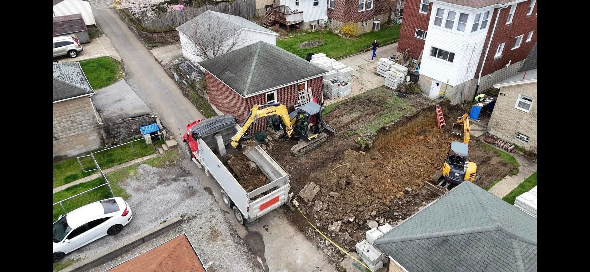 Aerial view of a construction site: an excavator loads dirt into a dump truck next to a residential house.