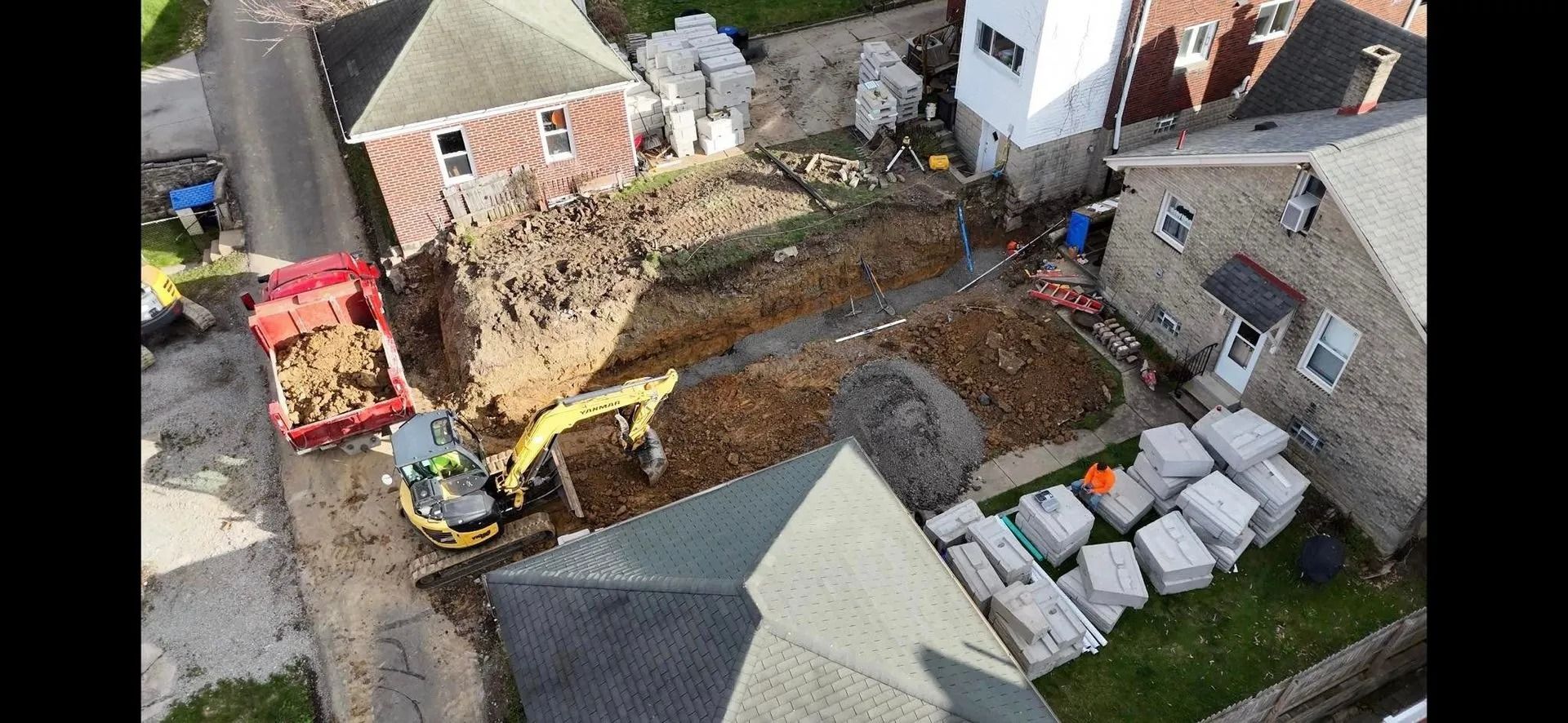 Excavator digging in yard next to houses, with a truck filled with dirt and stacks of concrete blocks.
