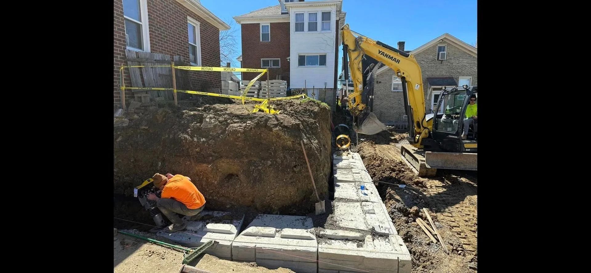 Construction site: excavator and worker building a retaining wall next to houses.