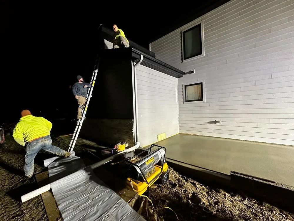 Construction workers at night. Men on roof and ladder, near new concrete patio and building with white siding.