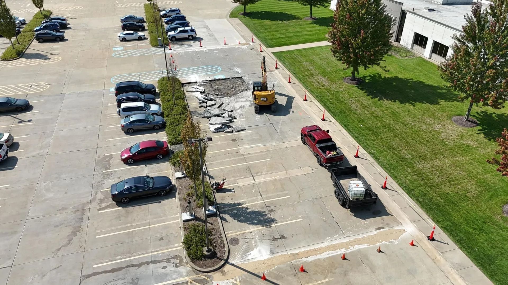 Aerial view of a parking lot under construction. An excavator and truck with a trailer are present.