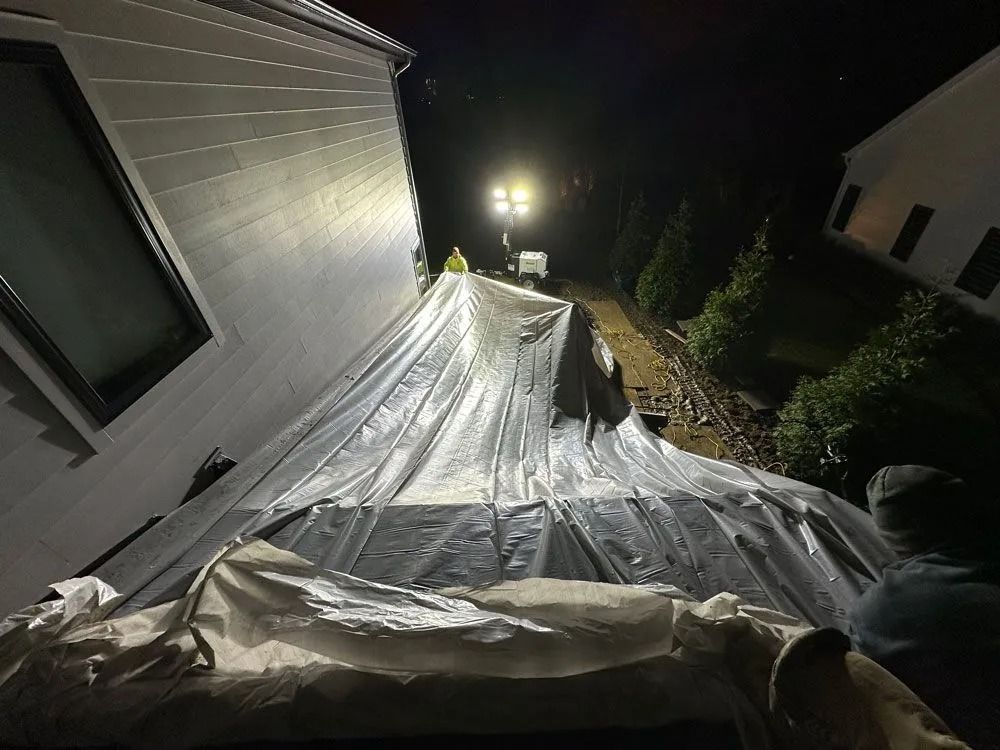 People covering a structure with a large tarp at night under bright lights. Building visible.