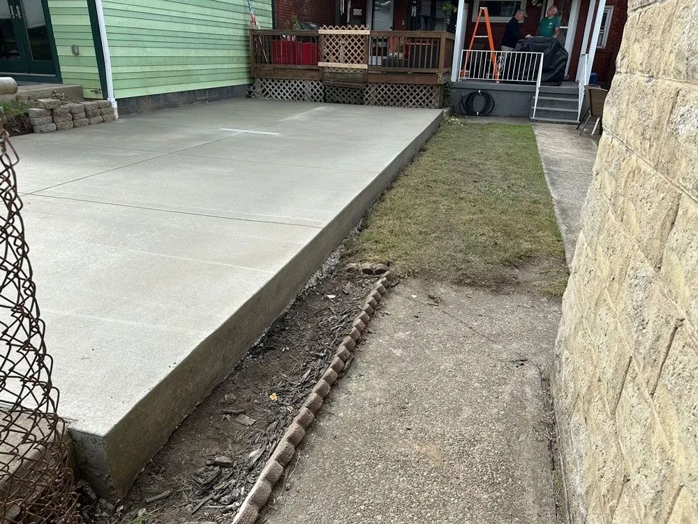 Concrete patio next to a narrow path with a border, beside a brick wall and grassy area.
