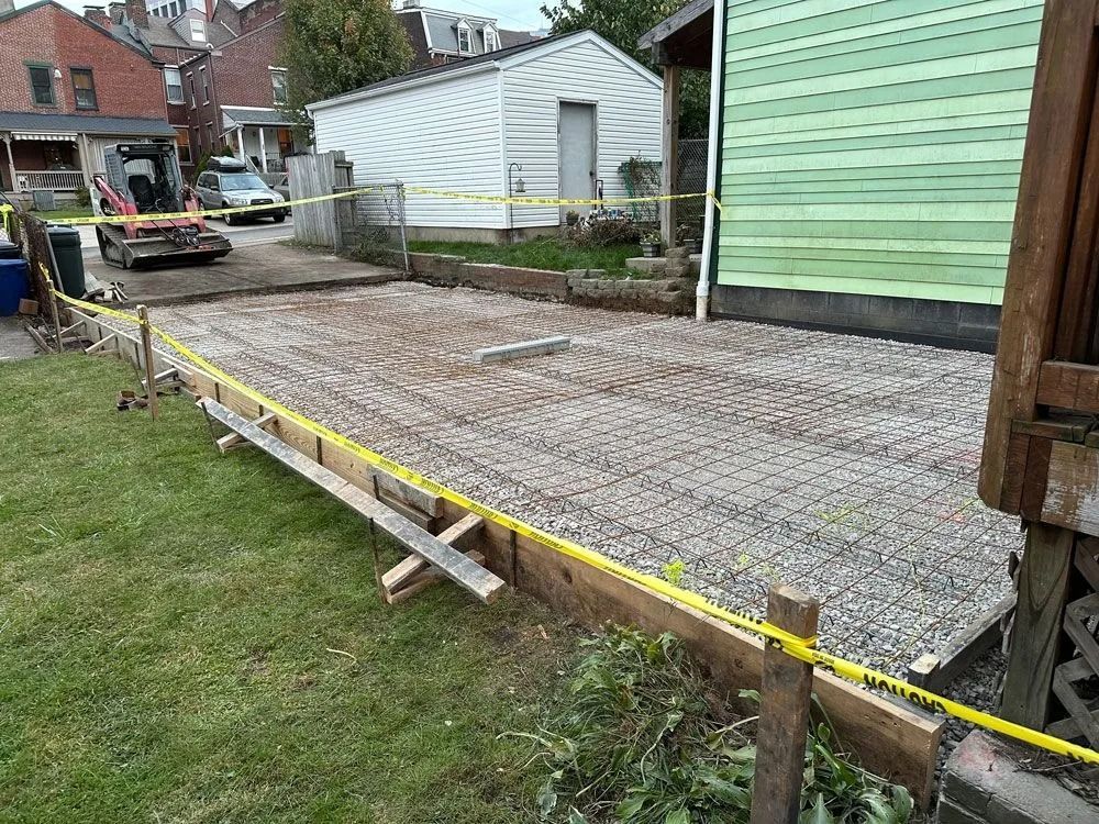 Construction site: concrete base with rebar grid, wooden frame, yellow caution tape, near a green building and yard.