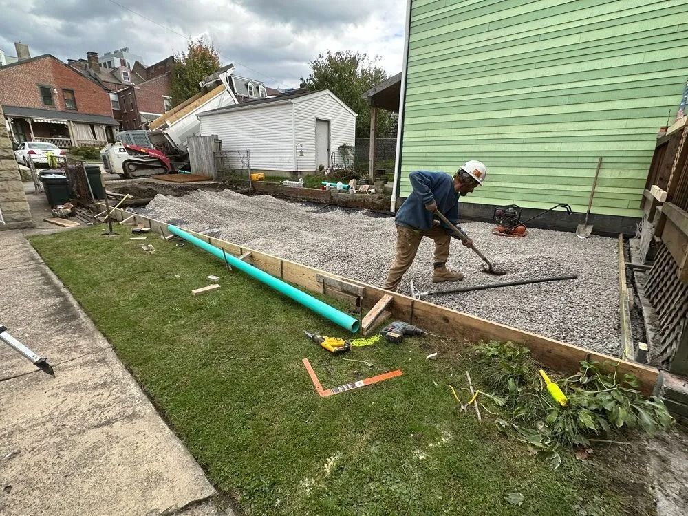 Construction worker leveling gravel, using a shovel, next to a newly formed concrete border and green grass.