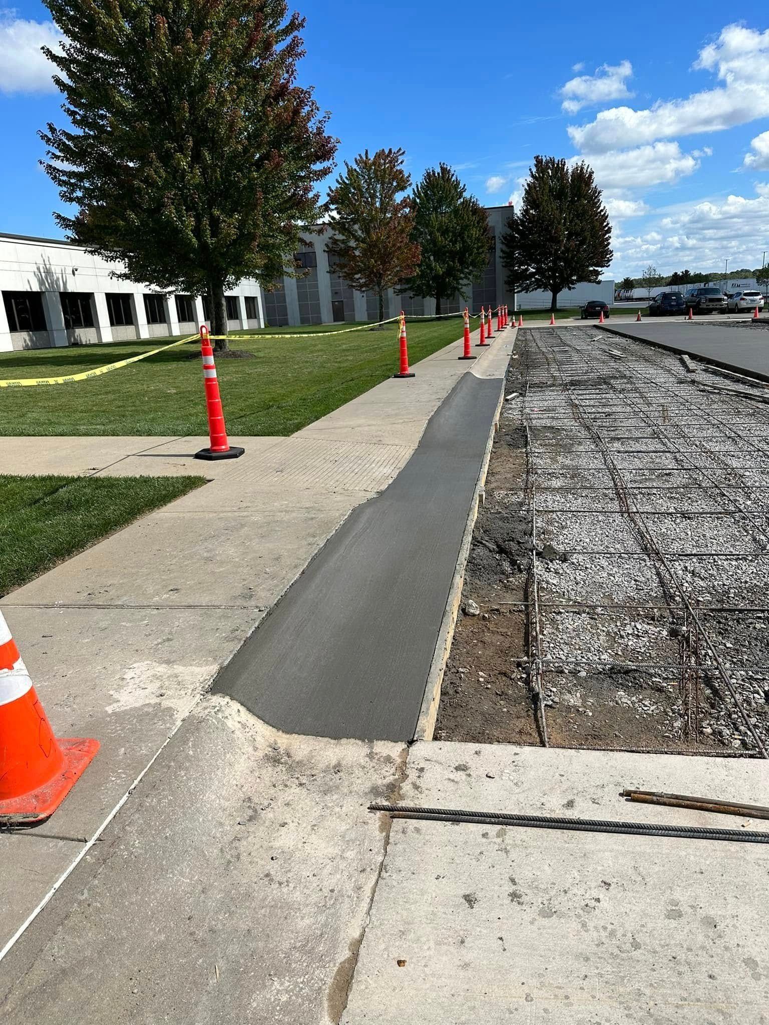 Sidewalk construction site with cones. Concrete path with a strip of new asphalt. Rebar and gravel are visible. Green grass and trees.