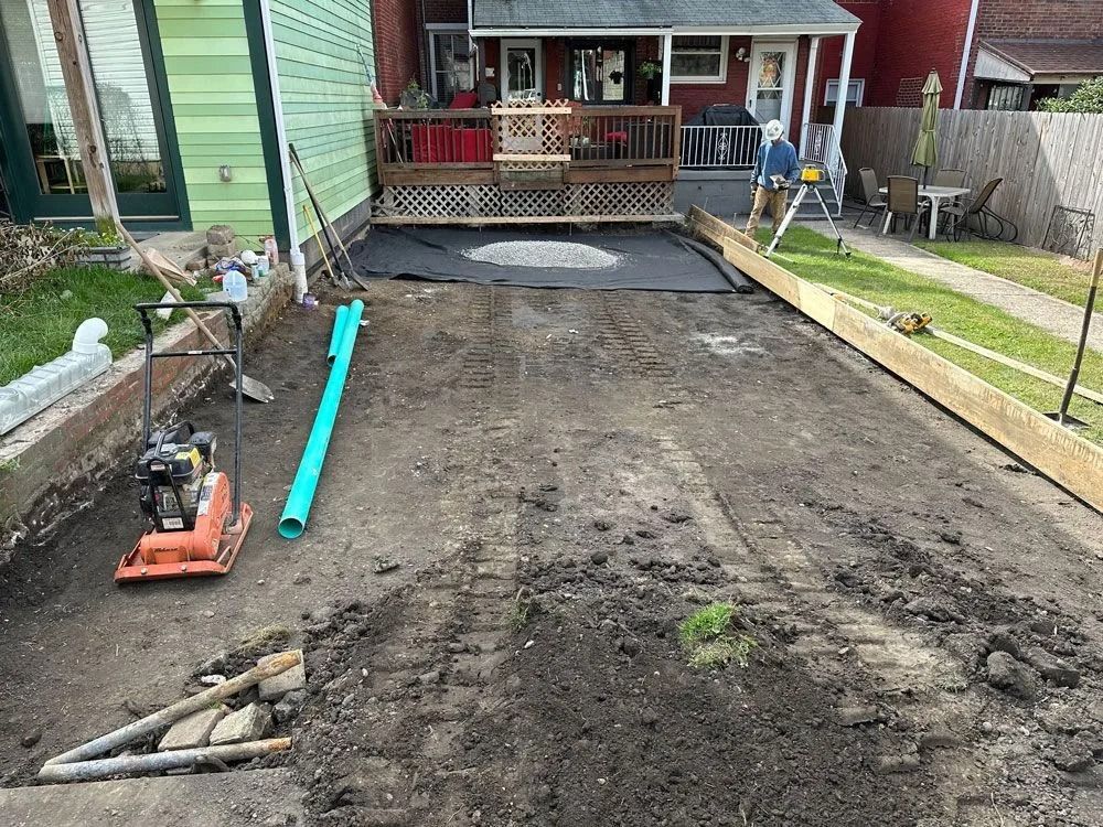 Backyard under construction; dirt, tools, new foundation, a deck in the background, green and brown tones.