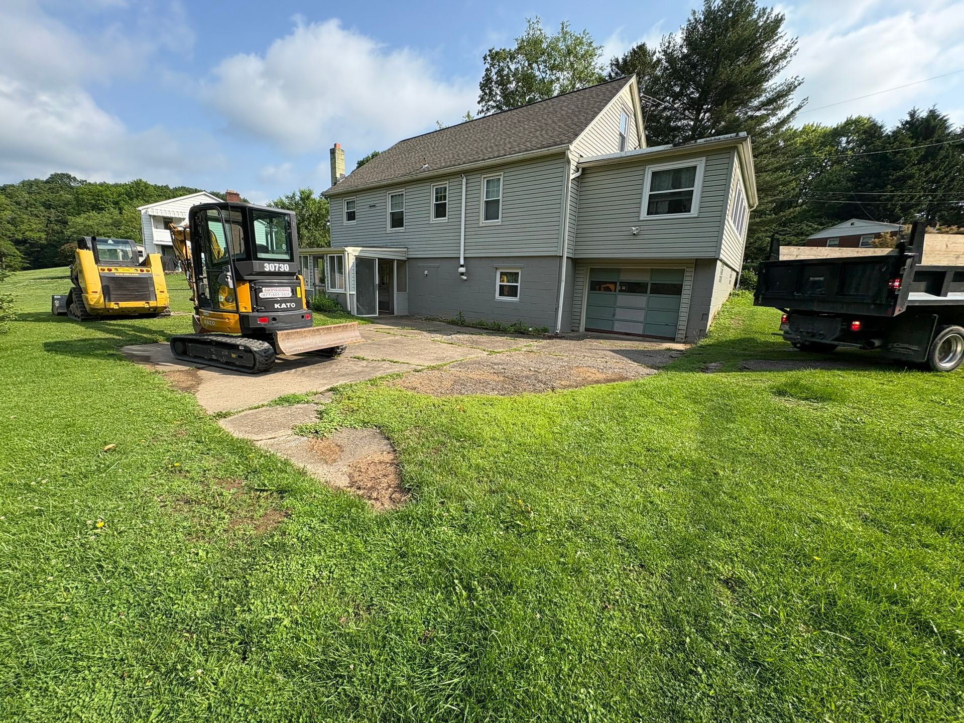 Construction site: mini excavator, dump truck, and skid steer in front of a house on a sunny day.