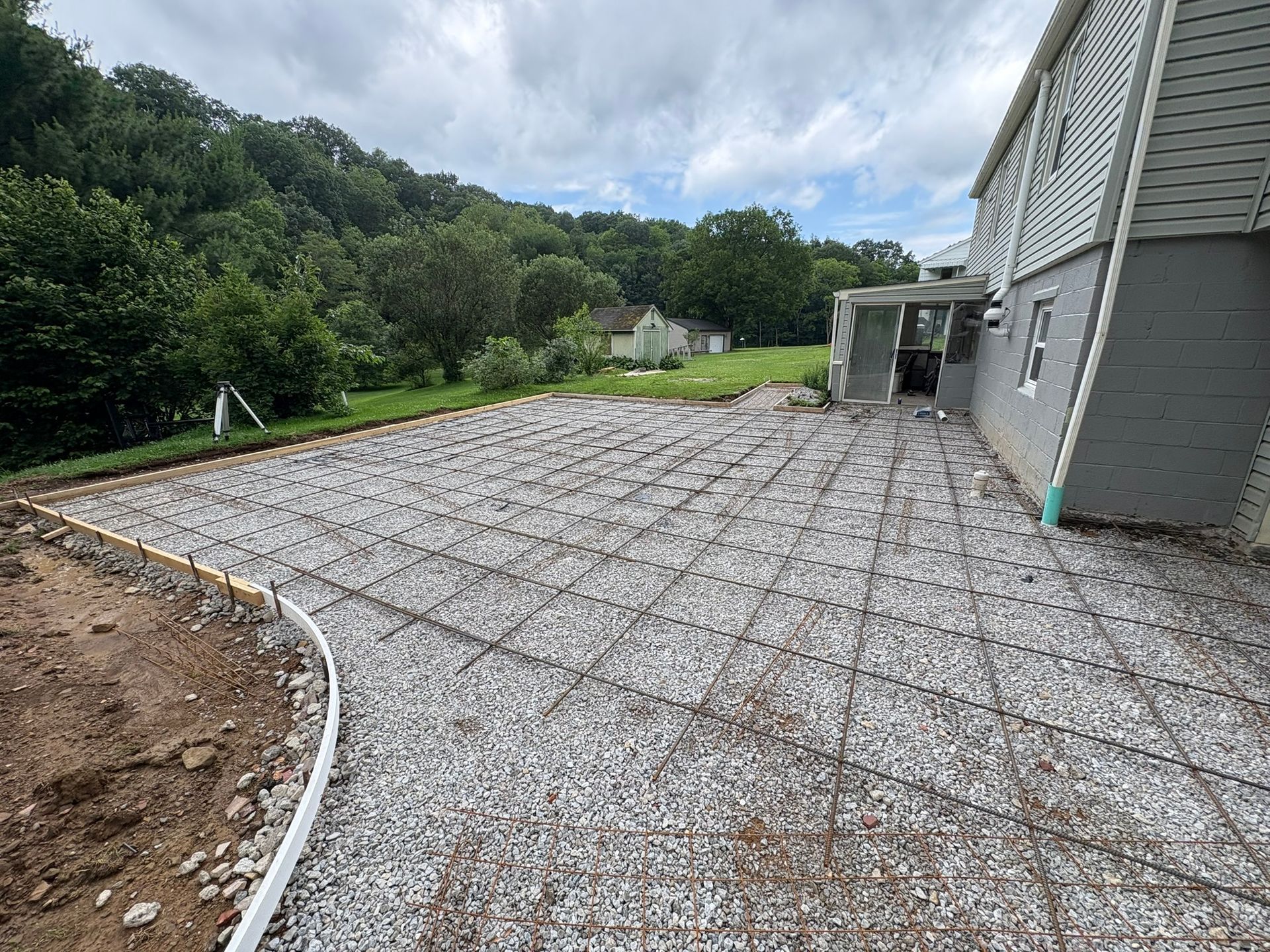 Gravel base with rebar grid prepared for a concrete patio next to a house under a cloudy sky.