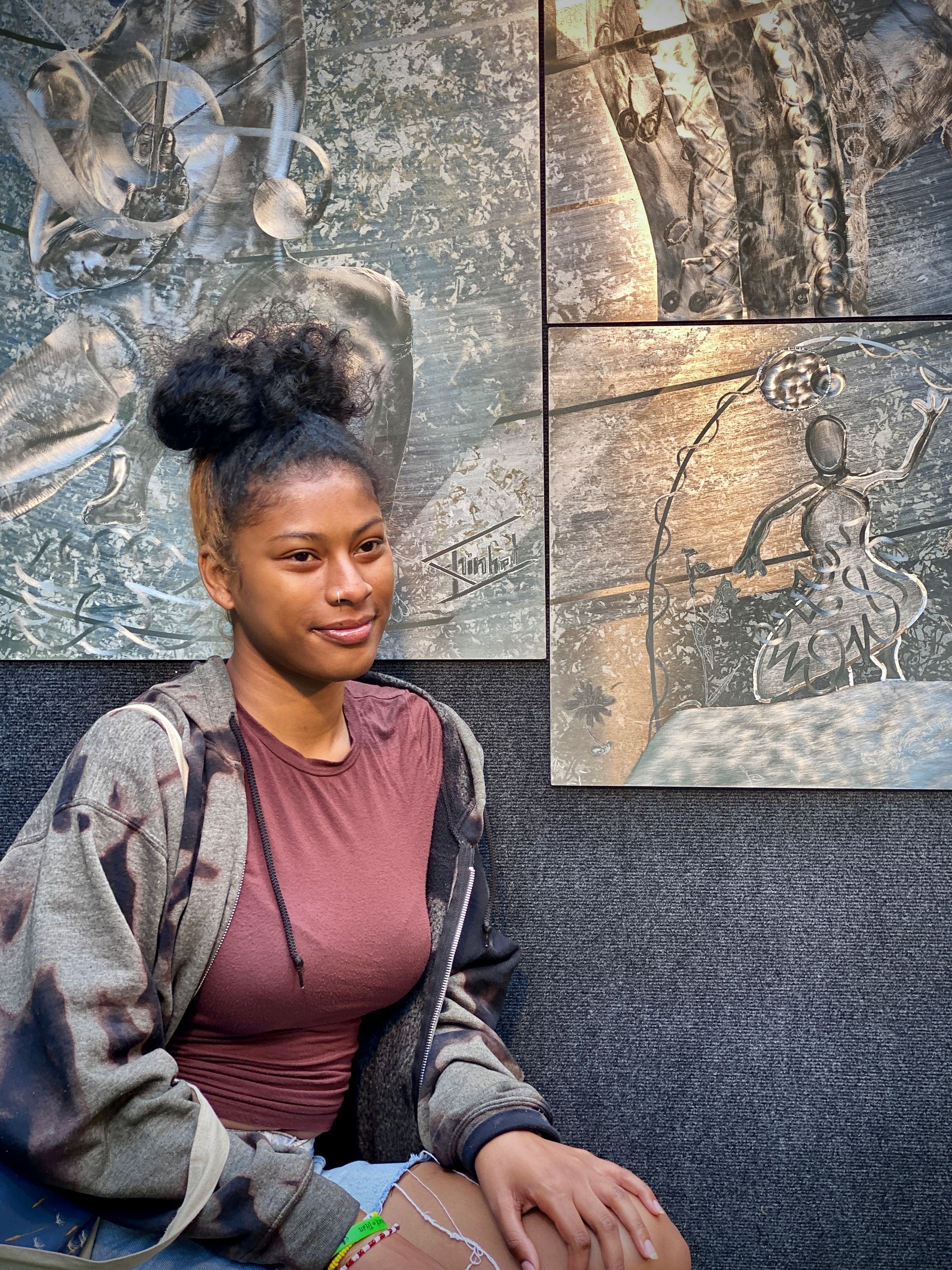 A woman is sitting in front of a painting on a wall.