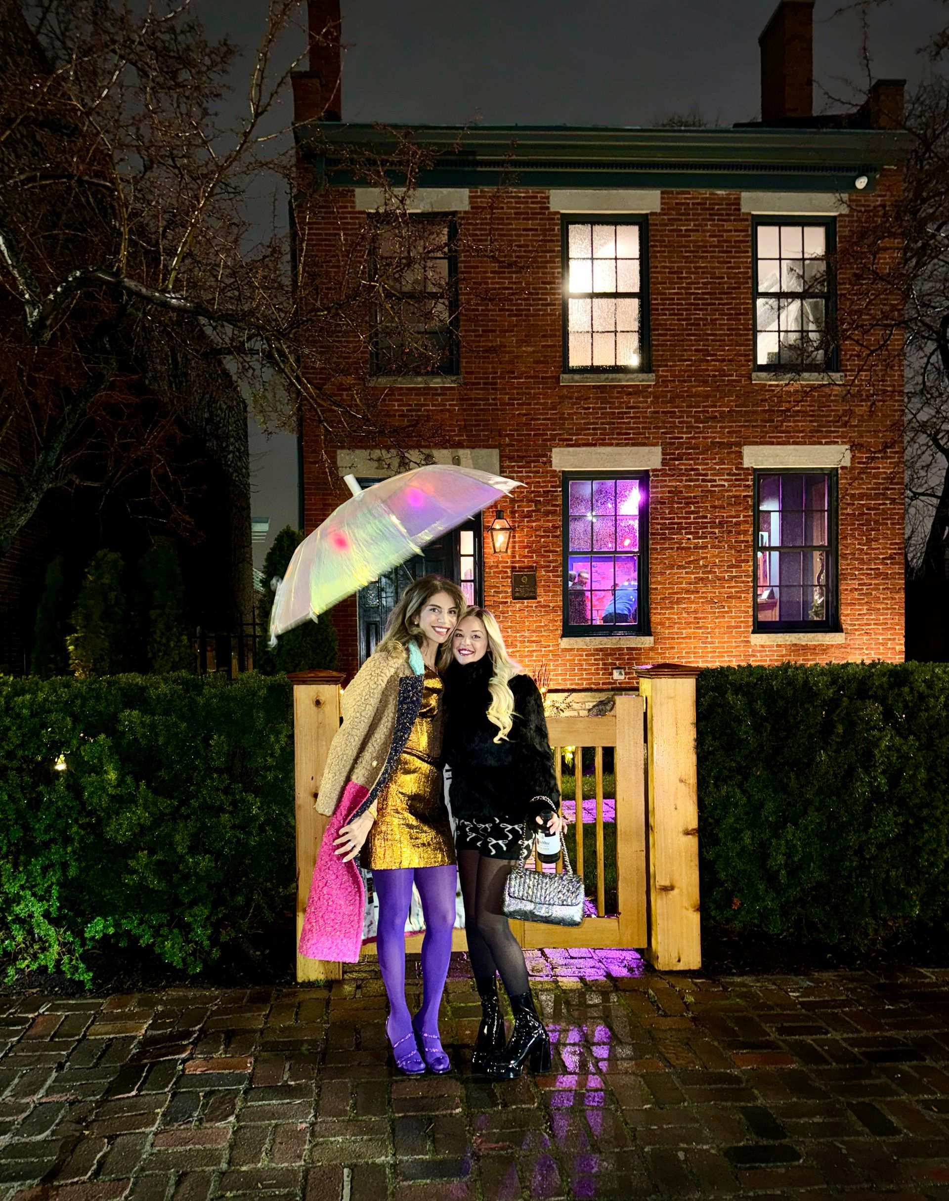Two women standing in front of a brick house holding an umbrella