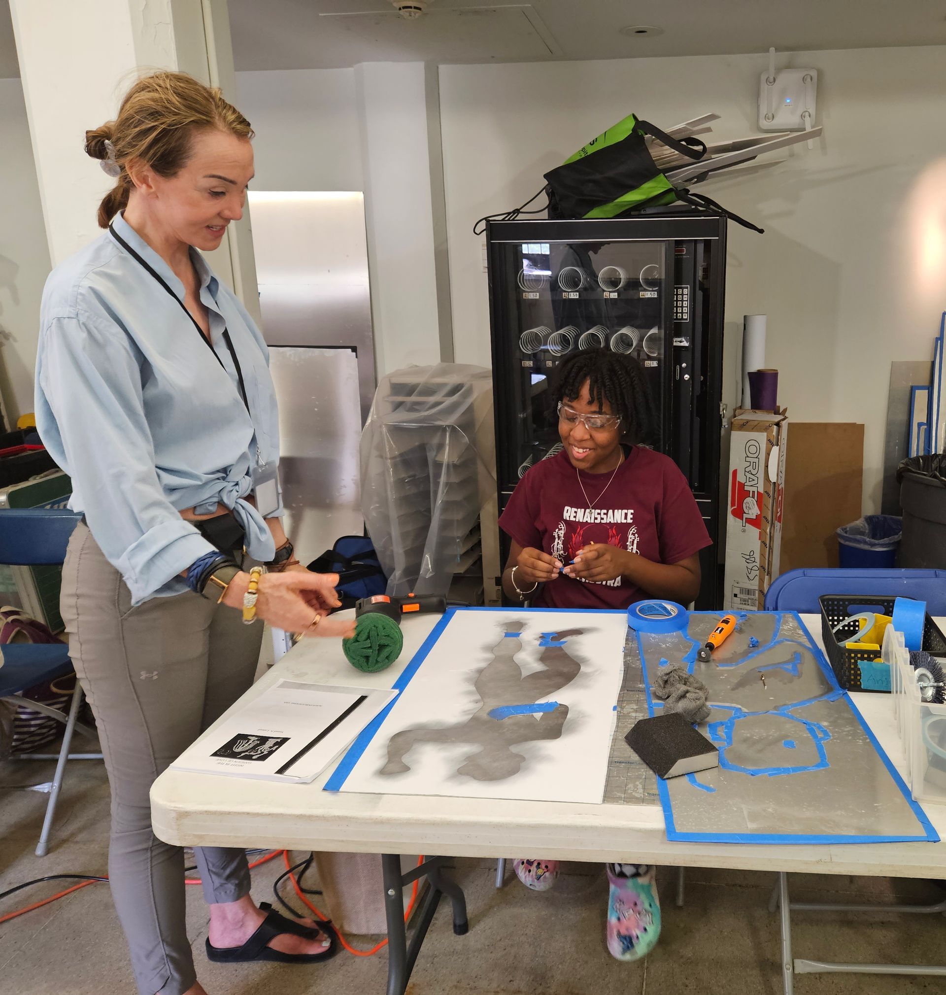 A girl in a burgundy shirt is sitting at a table with a woman standing next to her