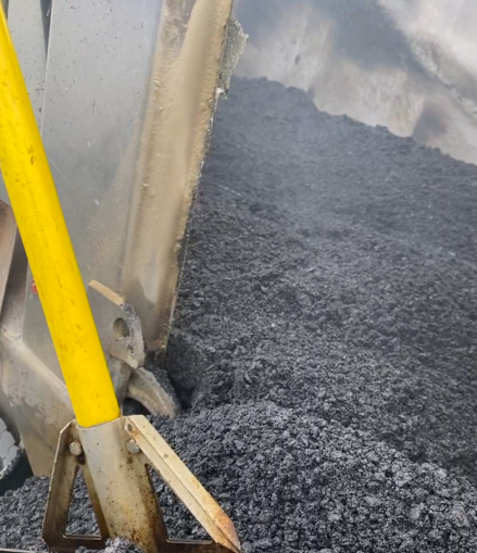 A yellow-handled shovel resting against the side of a metal dump truck bed filled with dark, coarse asphalt gravel.