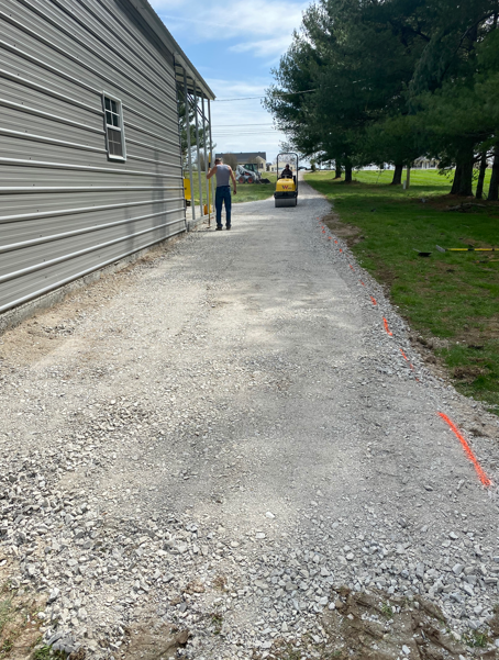 A gravel path being compacted by a small roller machine next to a metal building and grassy area.