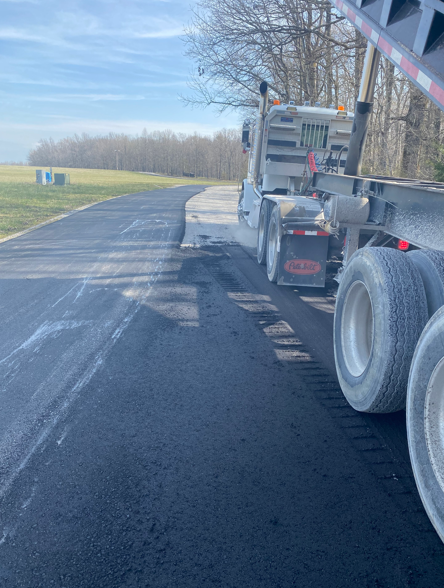 A white dump truck sits on the edge of a newly paved black asphalt road next to a field under a clear blue sky.