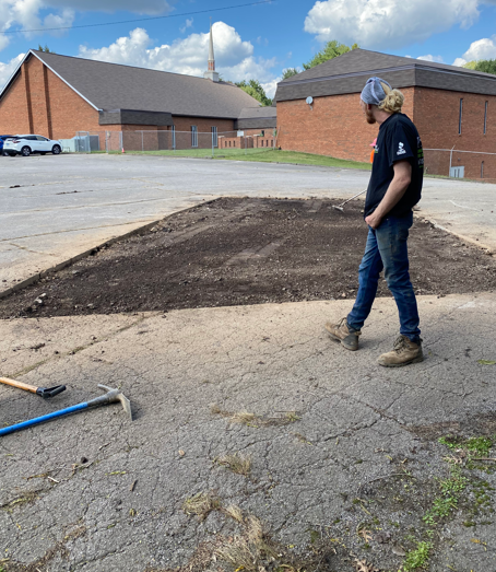 A person in a black shirt and jeans stands next to a rectangular patch of dirt in an outdoor paved parking lot.