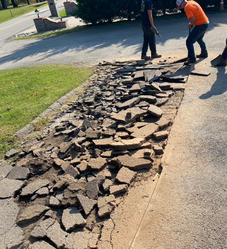 Two workers use tools to break up a damaged asphalt driveway.
