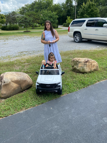 A person stands on grass behind a small child sitting in a white toy Chevrolet truck, near a white SUV and two large rocks.