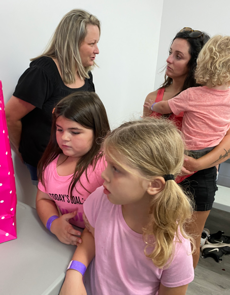 Two women and three children gathered in a room, with the adults watching as the children look toward a pink gift bag.